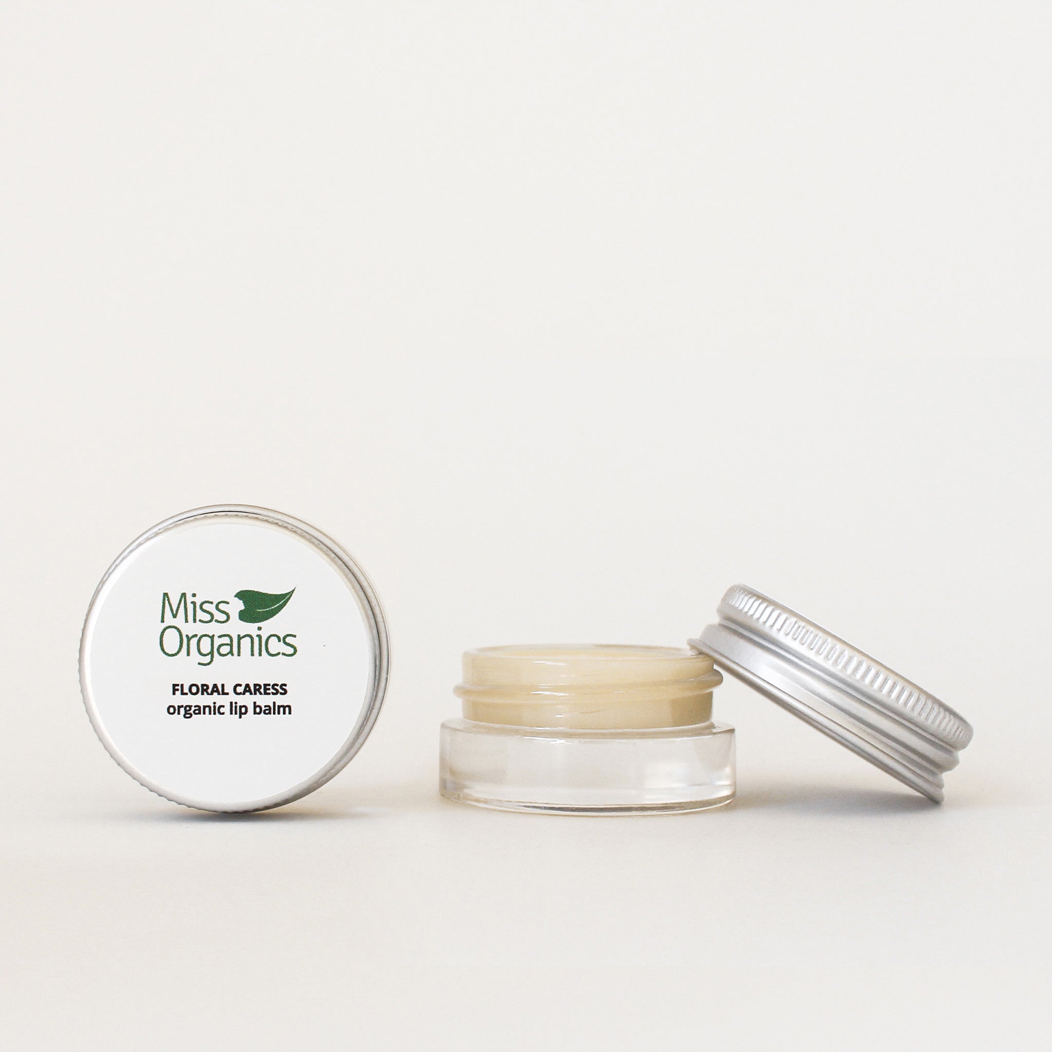 Floral Caress organic lip balm in glass pot with aluminium lid resting on pot on plain cream background